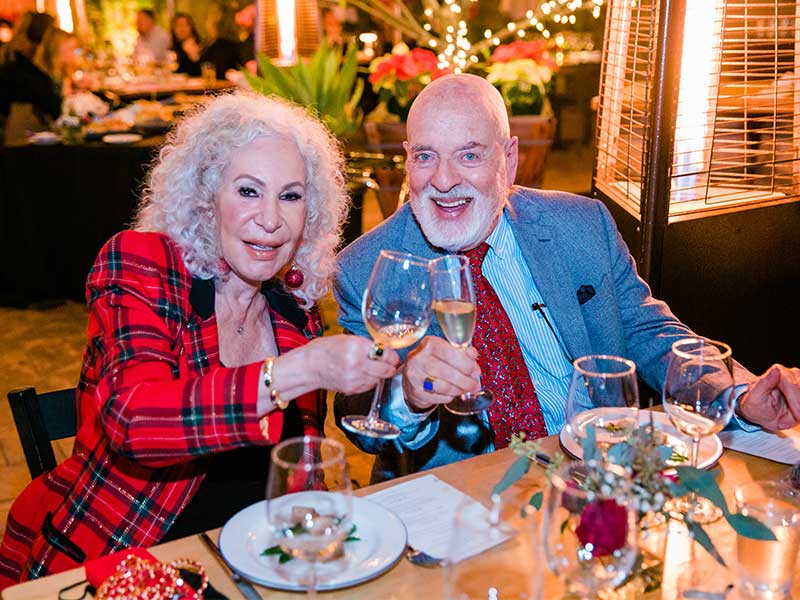 man and woman at a dining table looking at the camera and toasting glasses