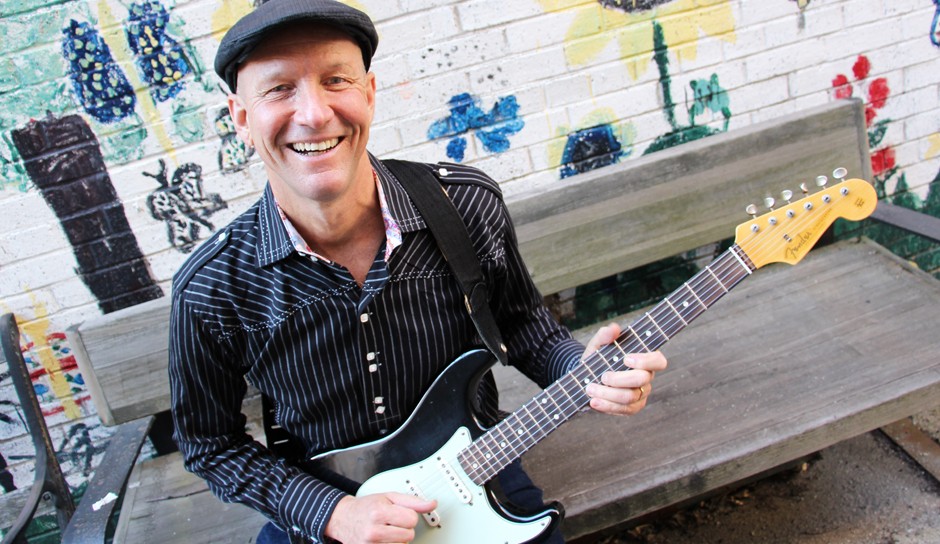 Man with beret in a black and white striped shirt sitting on a bench with an electric guitar