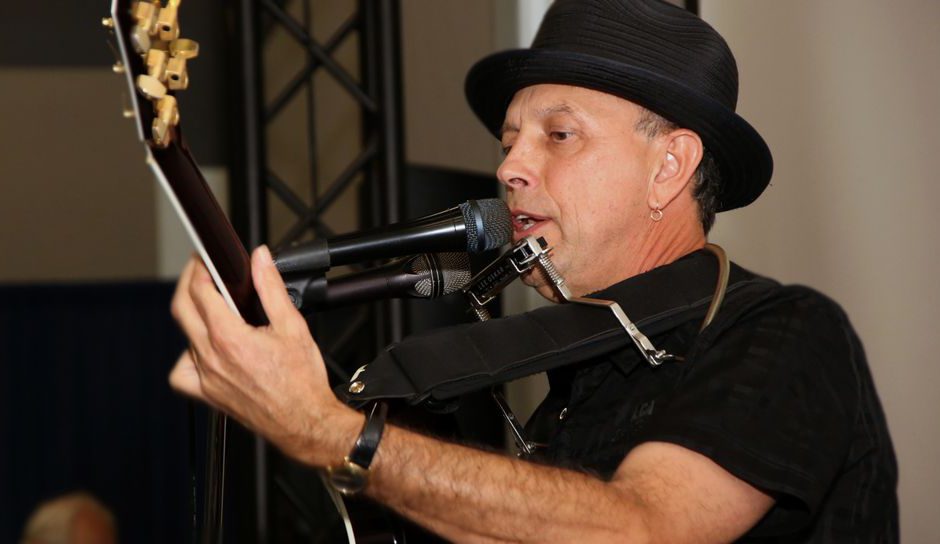 Man playing guitar signing into a mic wearing a black hat and black shirt