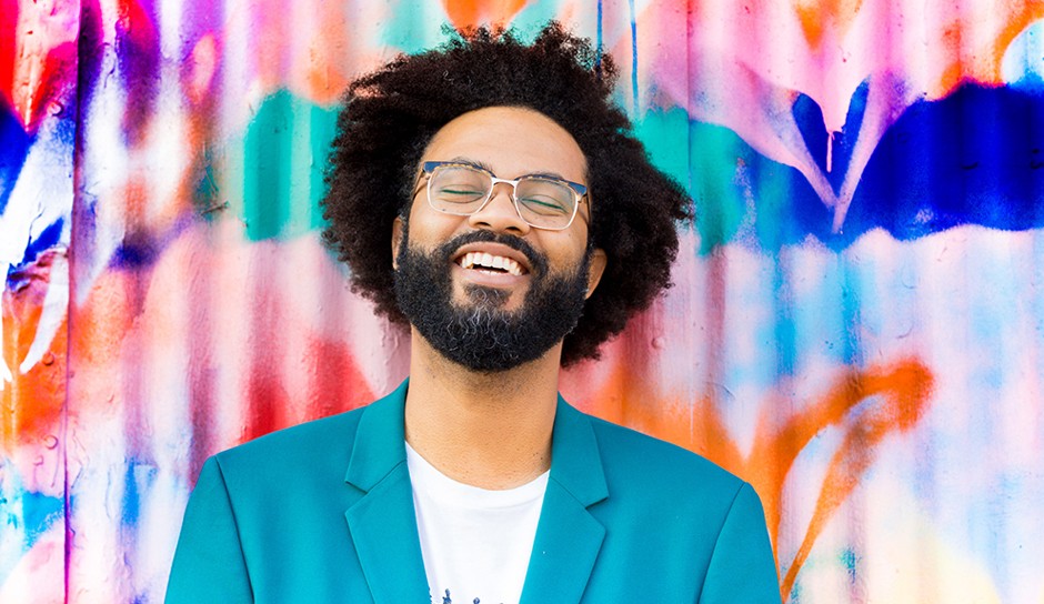 Man with black frizzy hair and beard in turquoise suit standing in front of a colorful painted wall