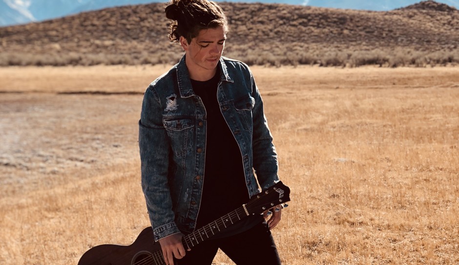 person standing in a dry grass field holding an acoustic guitar with a hill in the background