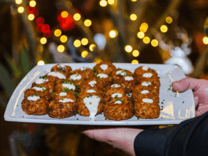 A hand holds a platter of round, breaded appetizers topped with white sauce and herbs, with festive lights behind.