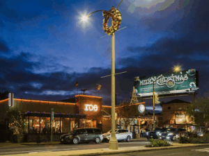 Street scene at dusk with holiday lights, a wreath on a lamp post, and a Merry Christmas billboard.